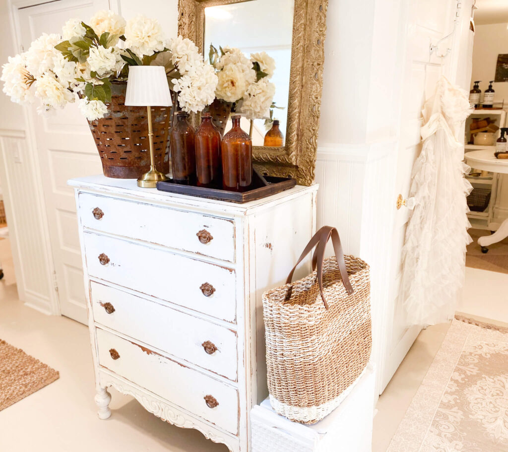 Image of white distressed vintage dresser with flowers, a lamp and antique amber bottles on top closer look at the side