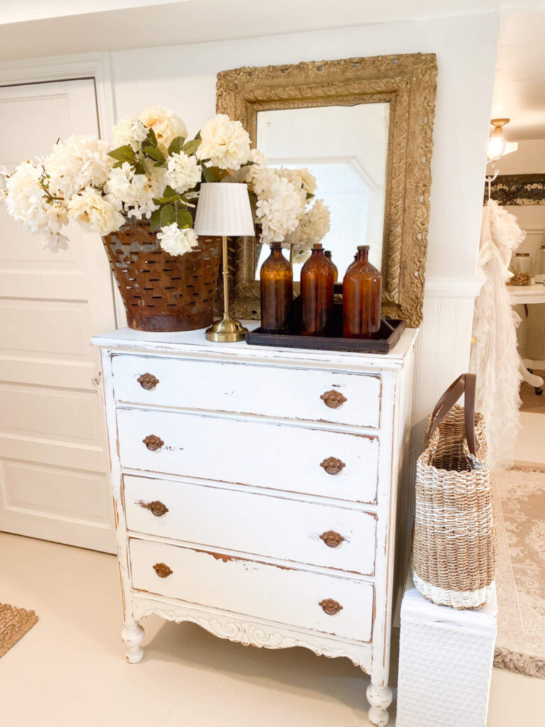 Image of white distressed vintage dresser with flowers, a lamp and antique amber bottles on top.