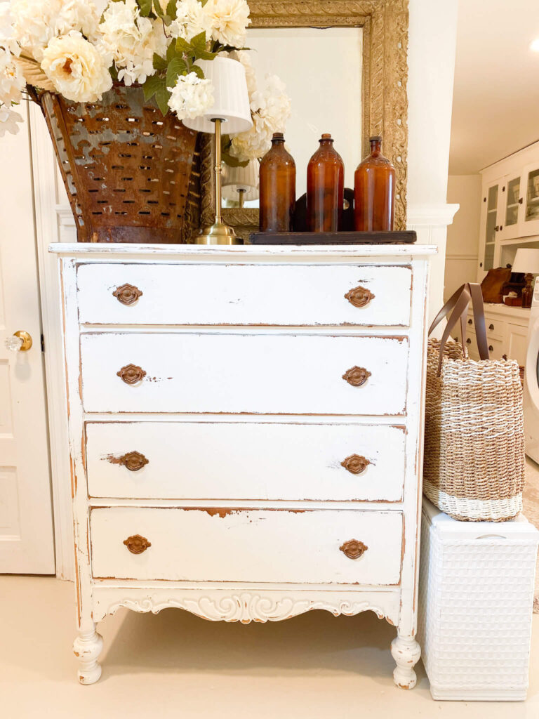 Image of white distressed vintage dresser with flowers, a lamp and antique amber bottles on top close up view