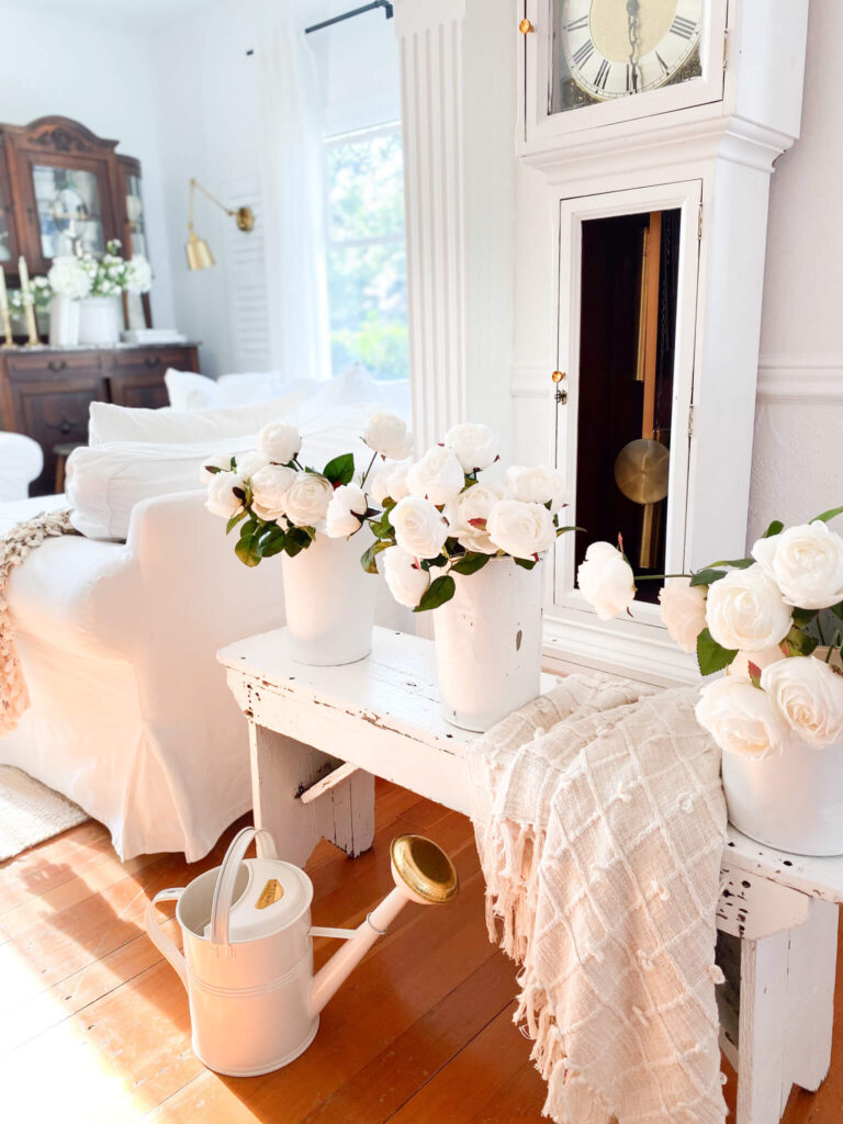 Close up side view of a small front entry with a vintage grandfather clock, antique white bench and 3 sap buckets of white roses