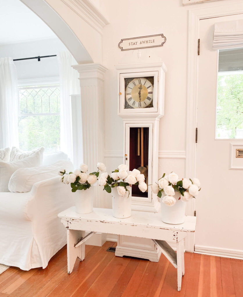 Small front entry with a grandfather clock, bench, white roses and Stay Awhile sign 