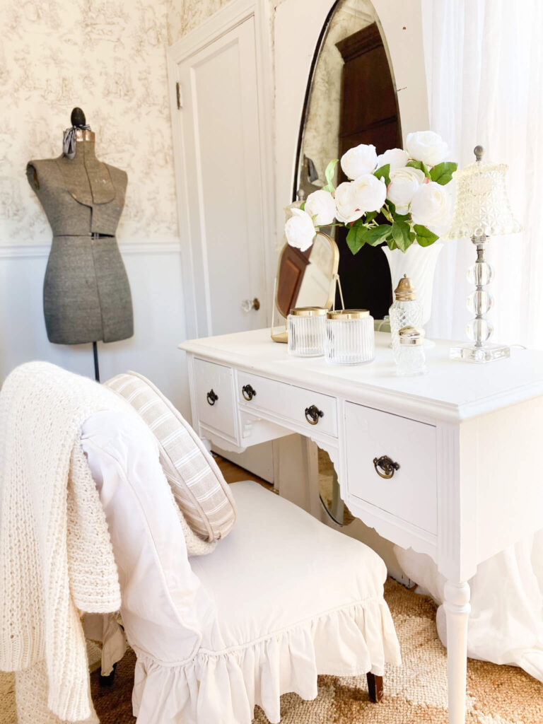 White antique desk with a vase of white roses, vintage glass lamps and an antique dress form in the background