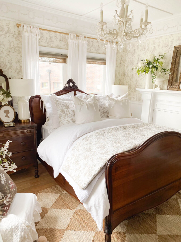 A cozy cottage bedroom with toile wallpaper, a white chandelier and white ruffled pillow shams