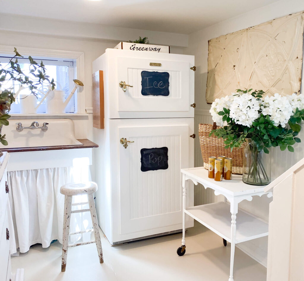Cottage basement laundry area with a white refrigerator with bead board panels, a farm sink and bar cart for serving drinks