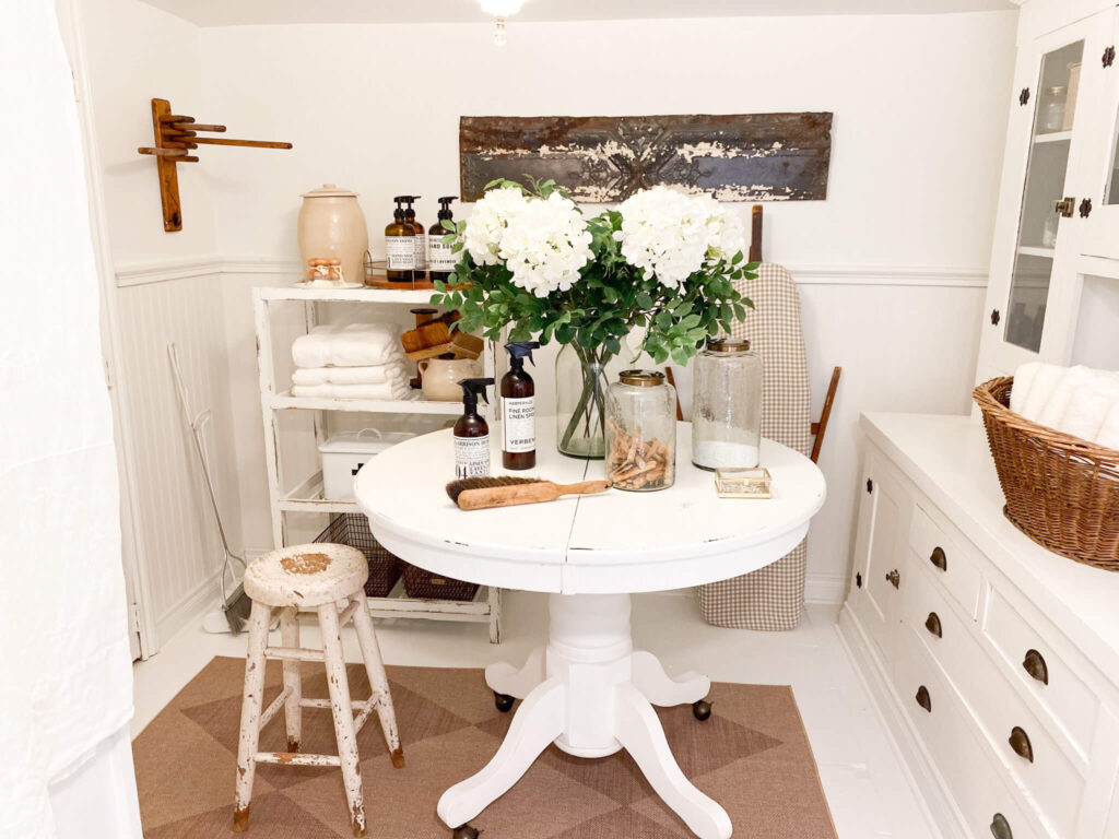 Small basement laundry room with a round antique table with a vase of flowers, an old wooden ironing board and vintage decor