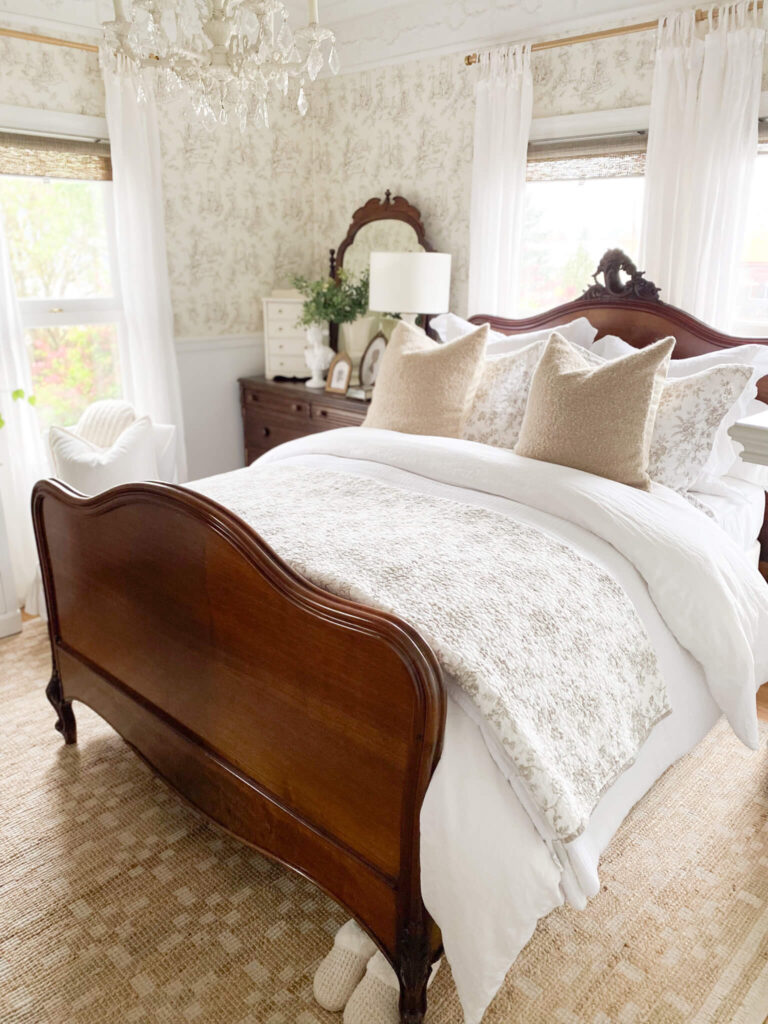 Cottage bedroom with antique bed, white comforter, textured taupe throw pillows and a toile quilt at the foot of the bed