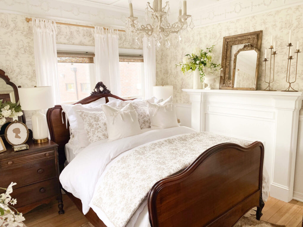 Image of an antique bed with light brown floral quilt, white ruffled pillow shams and an antique chandelier over the bed