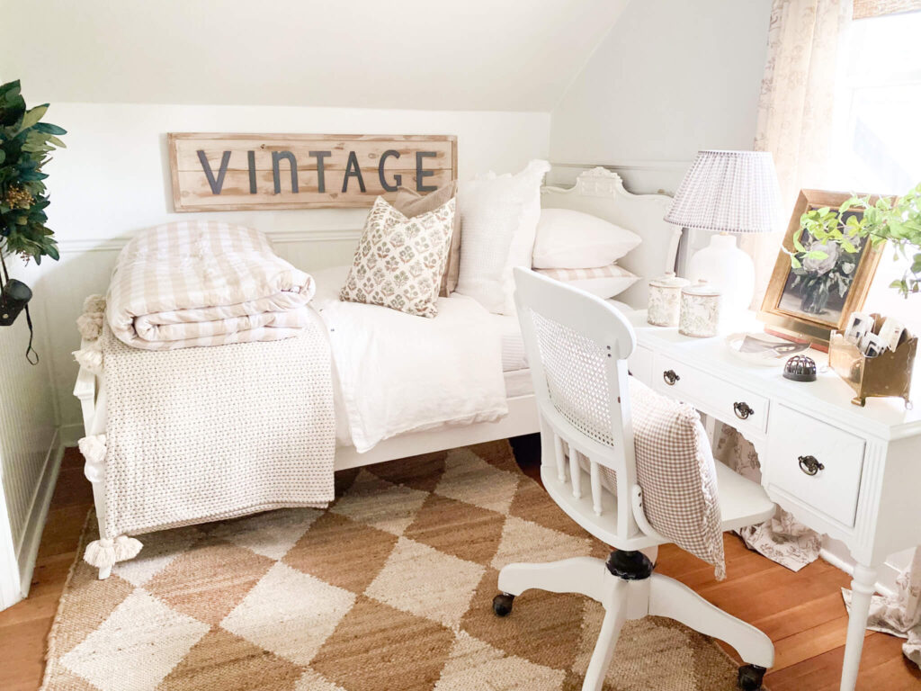A cozy bed in a white cottage bedroom with a vintage sign and a small antique desk and chair