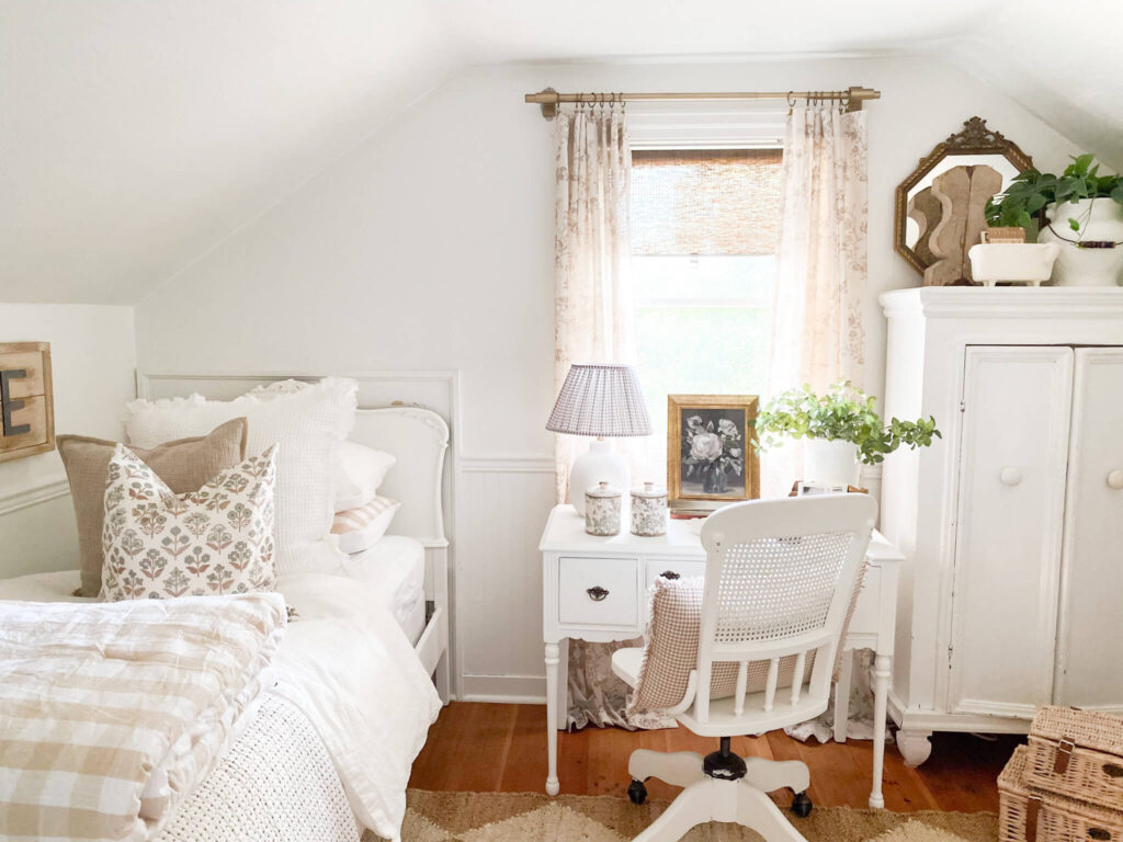 A cottage style bedroom with floral curtains, a small desk and chair and layered bedding with a nuetral gingham quilt