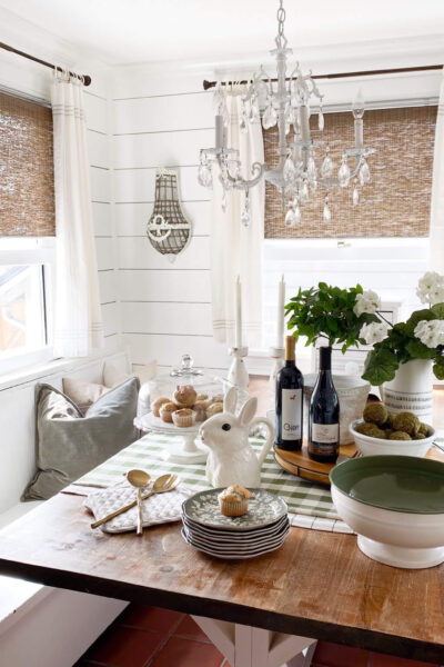 Image of a spring kitchen nook with a farmhouse table, and white geranium flowers, muffins, bunny pitcher and a green and white gingham tablecloth