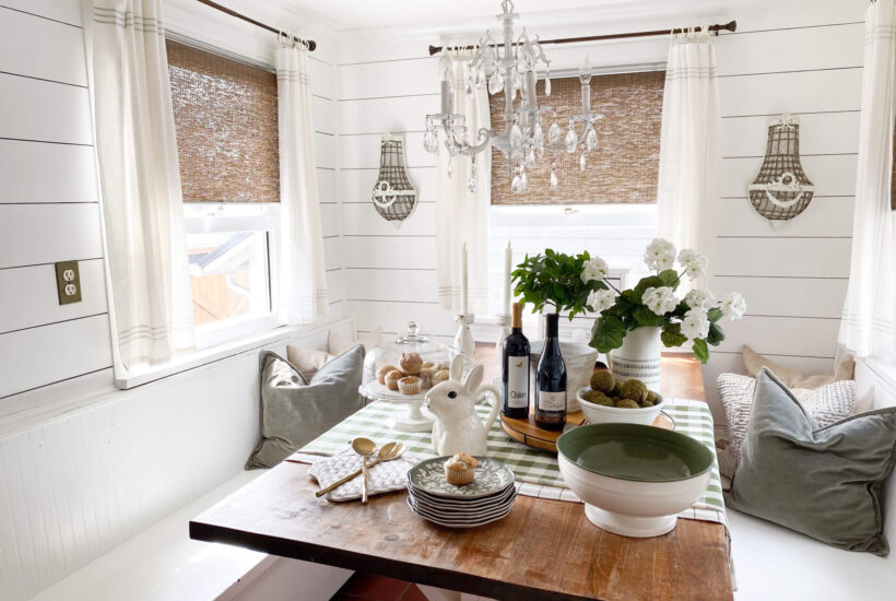 Image of a spring kitchen nook with a farmhouse table, and white geranium flowers, muffins, bunny pitcher and a green and white gingham tablecloth