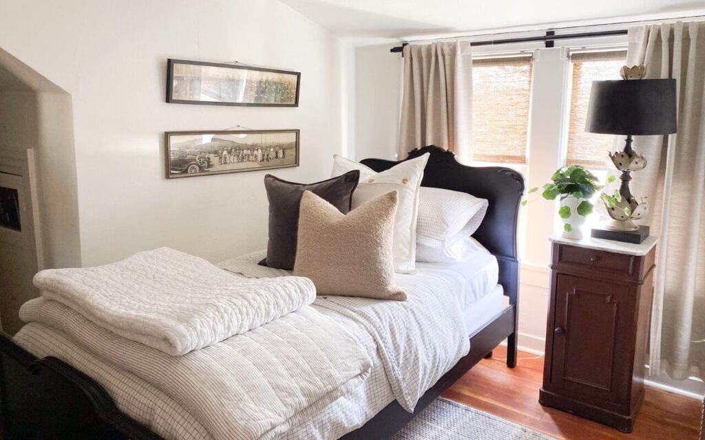 Small cottage bedroom with cozy white quilts and pillows with an antique nightstand and large vintage style lamp