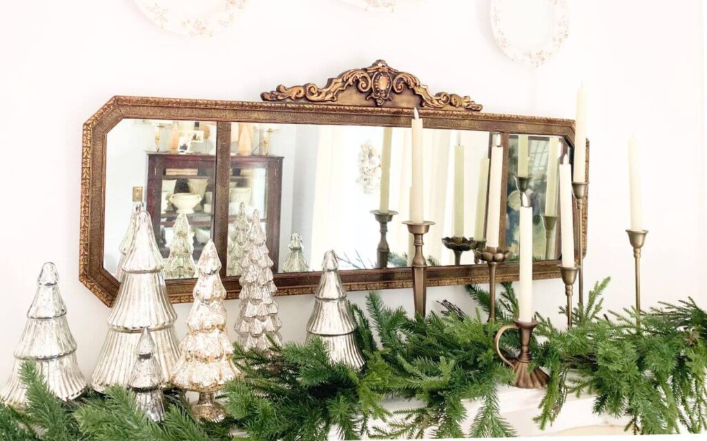 Vintage brass mirror hanging above a white mantel styled with christmas garland, a collection of mercury glass trees and many brass candelabras.