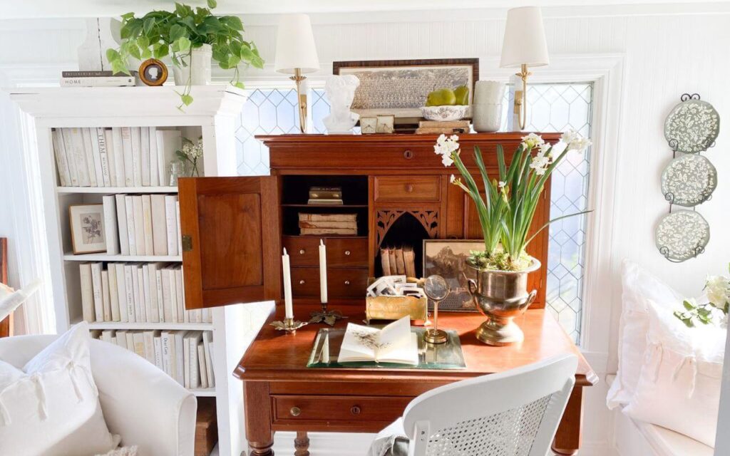 Antique desk in a sunroom home office surrounded by books and cottage-style decor