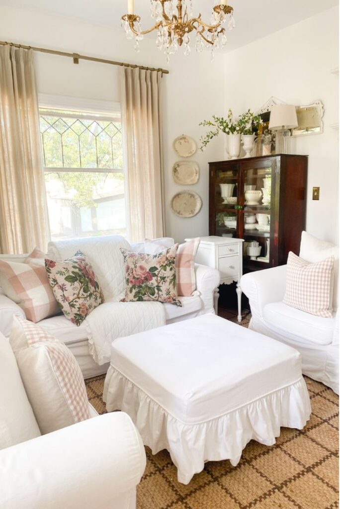 Cottage living room with pink gingham pillows and antique cabinet filled with white ironstone and white slipcovered furniture
